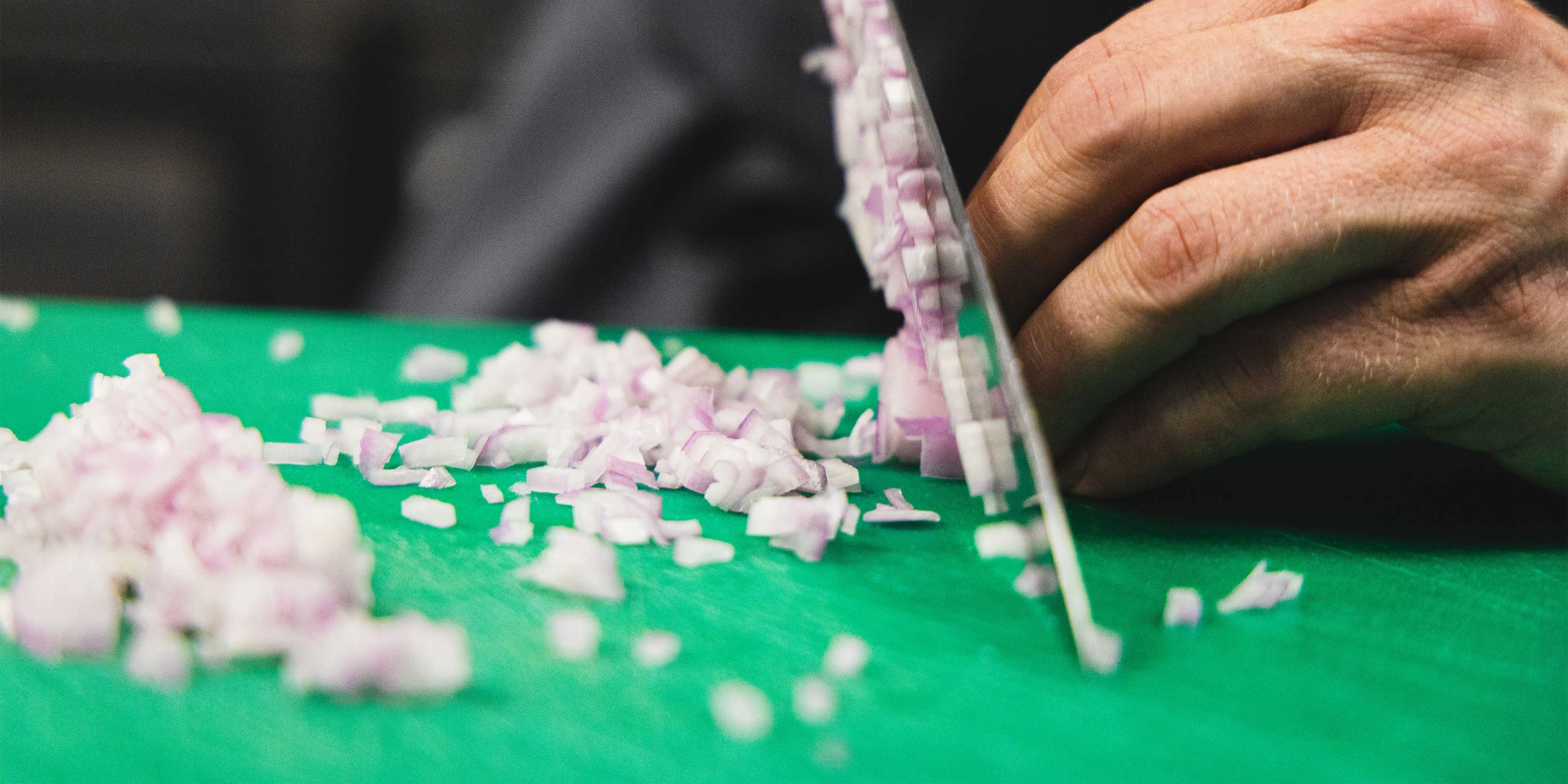 Close up of chef chopping onions with a knife on a green chopping board.
